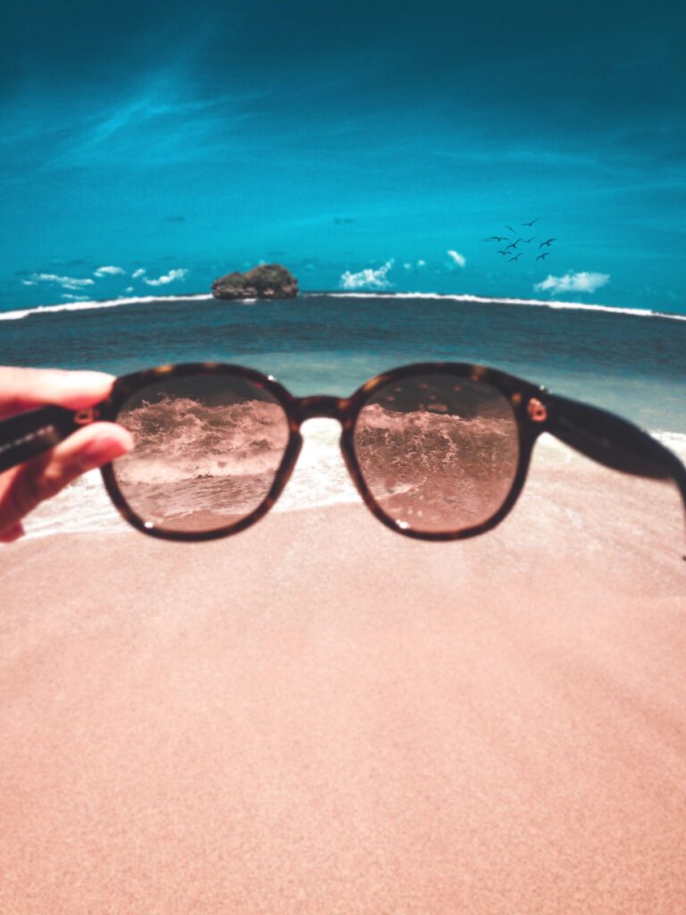Holding sunglasses up to a beach view, a reminder to switch off from work and find perspective on holiday.